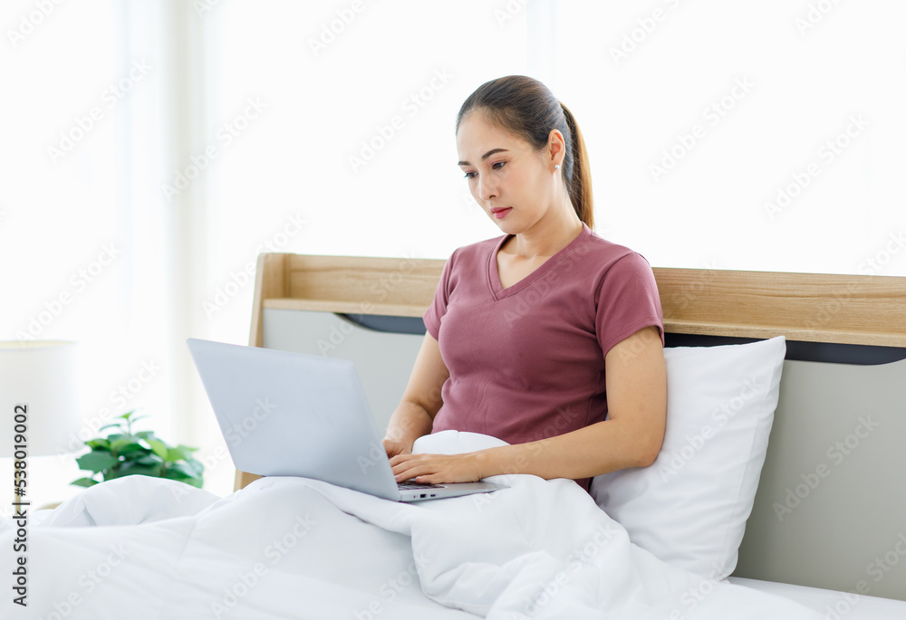 Millennial Asian young happy cheerful female housewife sitting smiling on bed and pillow under thick warm blanket using laptop notebook computer browsing surfing internet shopping online in bedroom Millennial Asian young happy cheerful female housewife sitting smiling on bed and pillow under thick warm blanket using laptop notebook computer browsing surfing internet shopping online in bedroom