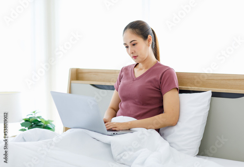 Millennial Asian young happy cheerful female housewife sitting smiling on bed and pillow under thick warm blanket using laptop notebook computer browsing surfing internet shopping online in bedroom