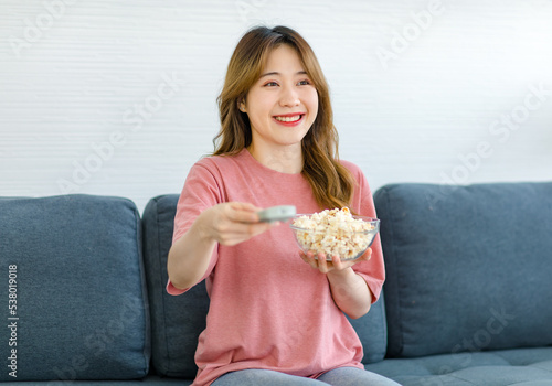 Millennial Asian young happy cheerful female housewife sitting smiling on cozy sofa holding popcorn snack glass bowl using remote control changing movie television channel in living room on weekend