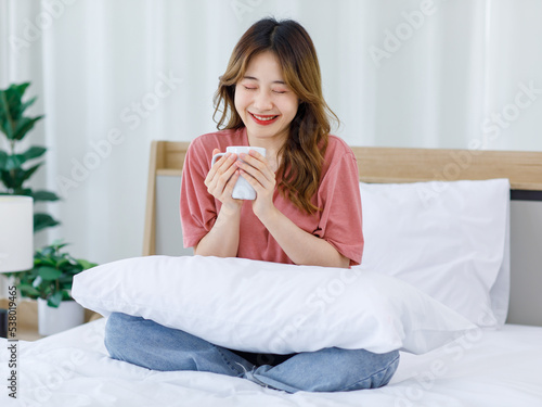Millennial Asian young happy cheerful female teenager sitting smiling on cozy bed in bedroom at home while drinking hot coffee in morning