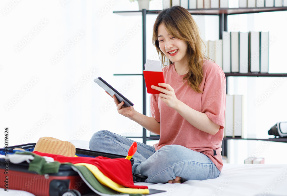 Millennial Asian young happy cheerful female teenager sitting smiling on bed in bedroom holding passport boarding pass air ticket while preparing packing clothes in trolley luggage for holiday trip Millennial Asian young happy cheerful female teenager sitting smiling on bed in bedroom holding passport boarding pass air ticket while preparing packing clothes in trolley luggage for holiday trip