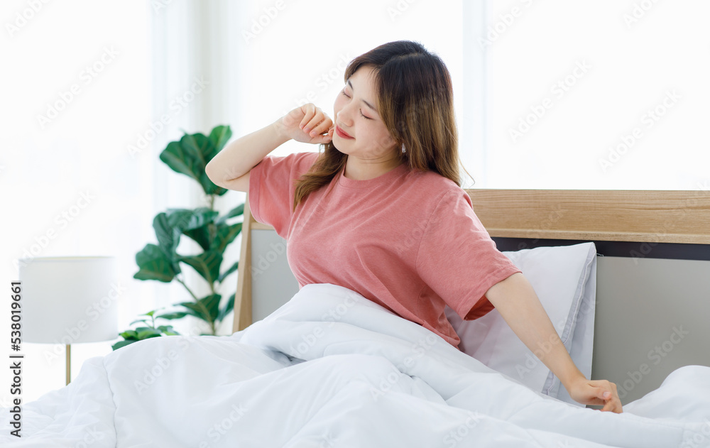 Millennial Asian young happy cheerful lazy sleepy female teenager sitting laying smiling on pillow under thick warm blanket on cozy bed in bedroom at home stretching arms after waking up in morning Millennial Asian young happy cheerful lazy sleepy female teenager sitting laying smiling on pillow under thick warm blanket on cozy bed in bedroom at home stretching arms after waking up in morning