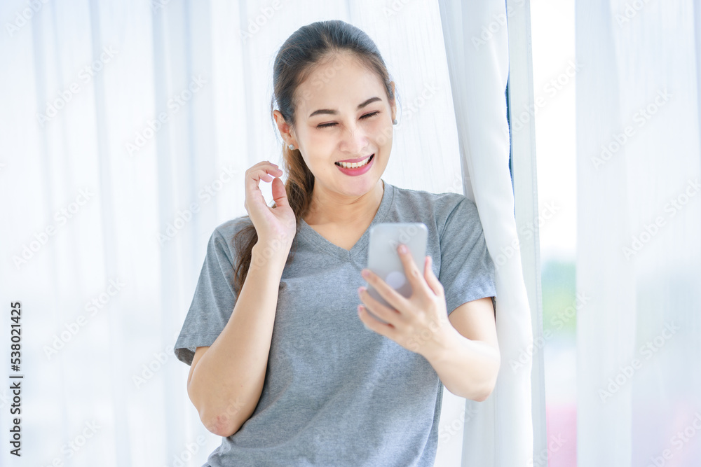 Millennial Asian young happy cheerful female housewife in casual outfit standing smiling using touchscreen smartphone video call chatting talking greeting with friends in front curtain background Millennial Asian young happy cheerful female housewife in casual outfit standing smiling using touchscreen smartphone video call chatting talking greeting with friends in front curtain background