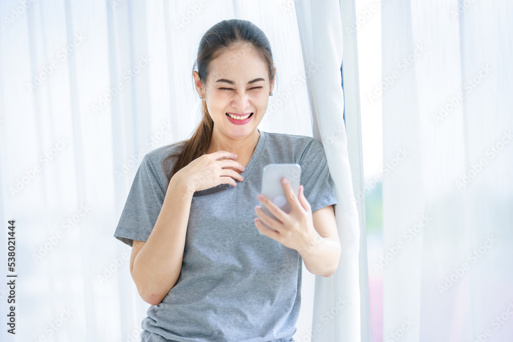 Millennial Asian young happy cheerful female housewife in casual outfit standing smiling using touchscreen smartphone video call chatting talking greeting with friends in front curtain background Millennial Asian young happy cheerful female housewife in casual outfit standing smiling using touchscreen smartphone video call chatting talking greeting with friends in front curtain background