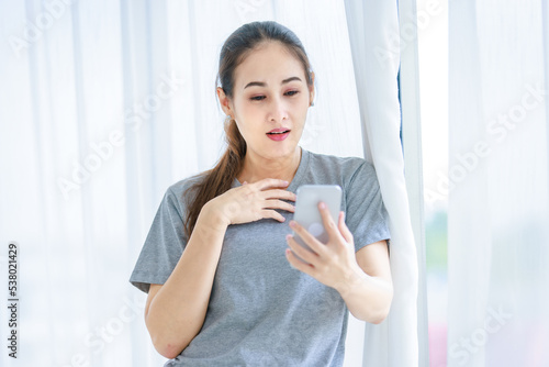 Free Download Millennial Asian young happy cheerful female housewife in casual outfit standing smiling using touchscreen smartphone video call chatting talking greeting with friends in front curtain background - GFXLoad