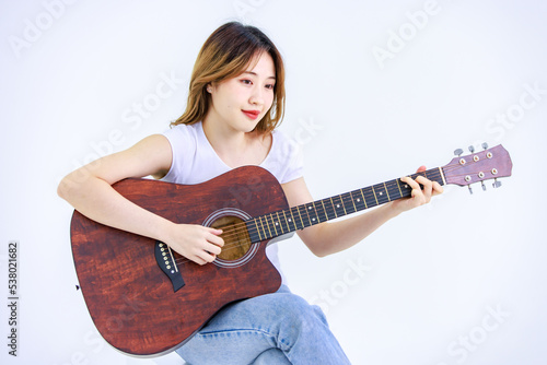 Isolated cutout studio full body shot of Millennial Asian young happy cheerful female teenager teen model in casual outfit sitting on square box smiling singing playing guitar on white background