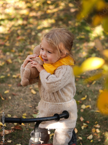 Portrait of cute little blond girl in yellow scarf hugging her toy teddy bear riding on bike outdoors in park