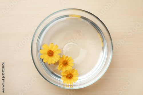 Glass bowl with water and yellow flowers on wooden table, top view