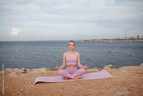 Young beautiful woman dressed pink sportwear  makes the healthy for body yoga exercises against the background of blue water of sea or ocean and sky