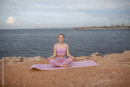Young beautiful woman dressed pink sportwear, makes the healthy for body yoga exercises against the background of blue water of sea or ocean and sky