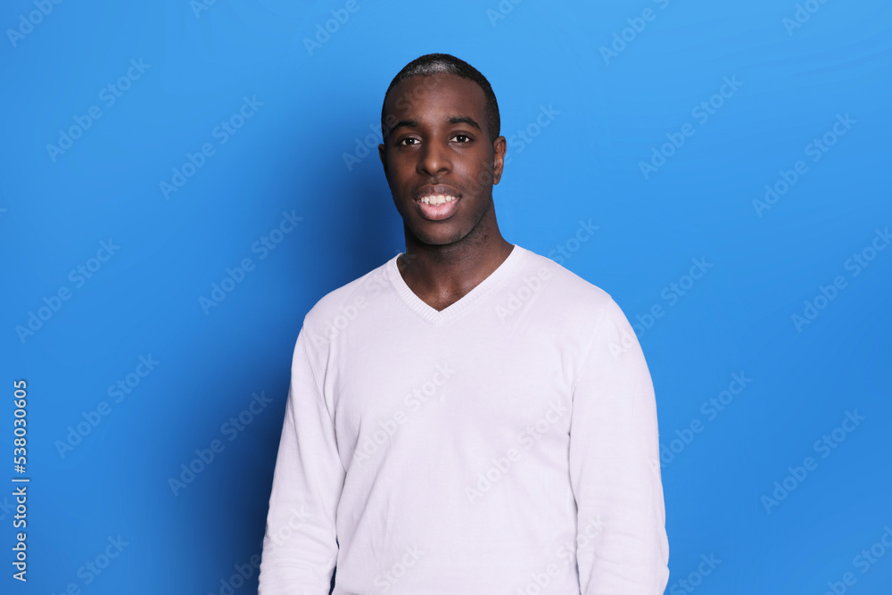 Smiling young African-american man in white casual sweater standing, looking at camera with wide toothy smile isolated on blue background Smiling young African-american man in white casual sweater standing, looking at camera with wide toothy smile isolated on blue background