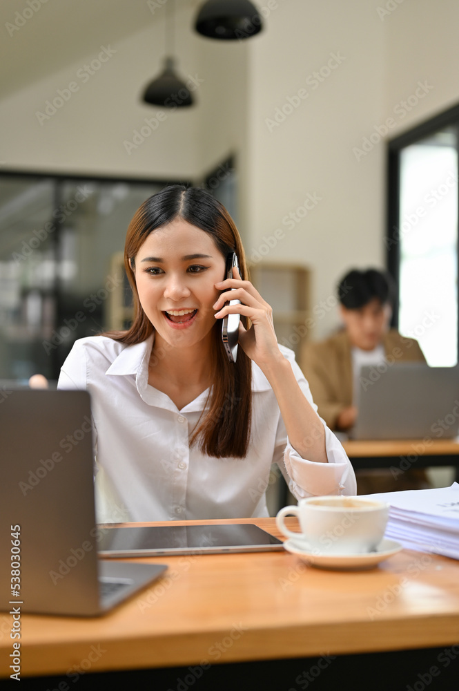Cheerful Asian female account executive is on the phone at her desk
