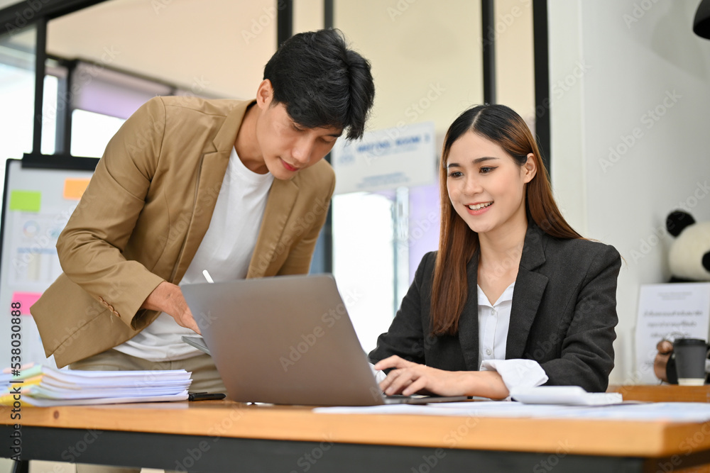Asian businesswoman working on her project on laptop while discussing with her male colleague Asian businesswoman working on her project on laptop while discussing with her male colleague