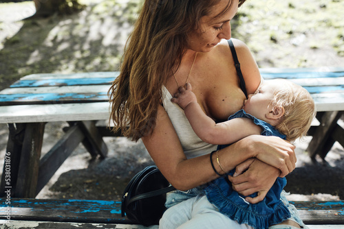 Mother feeding breast milk to toddler daughter while sitting on bench photo