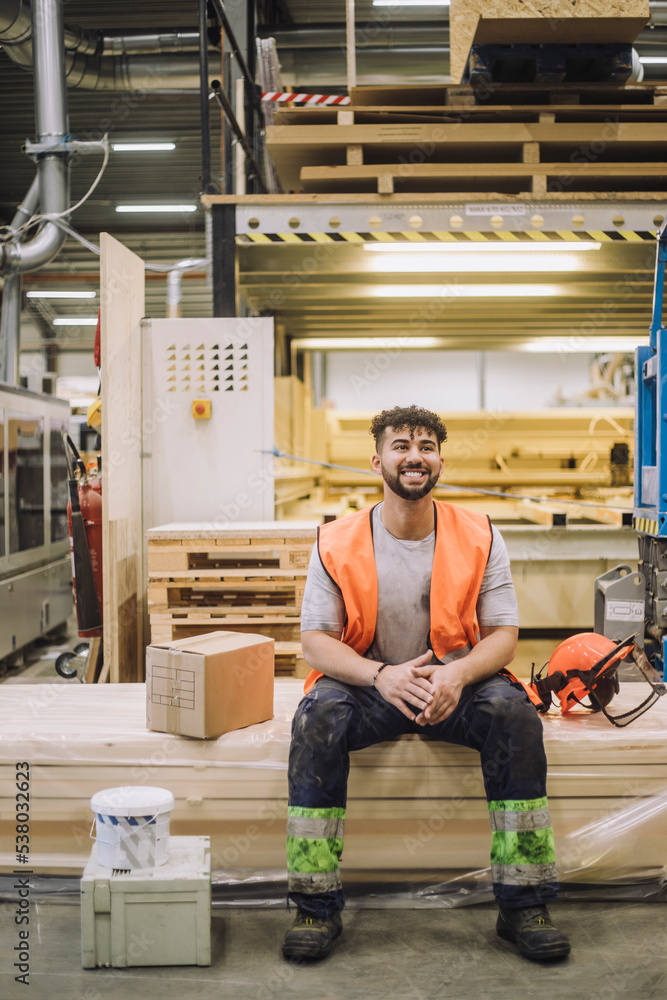 Full length of young carpenter sitting by hardhat in warehouse while looking away Full length of young carpenter sitting by hardhat in warehouse while looking away