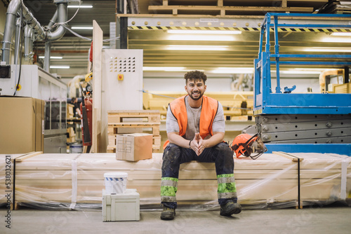 Portrait of smiling young blue-collar worker sitting by hardhat in warehouse