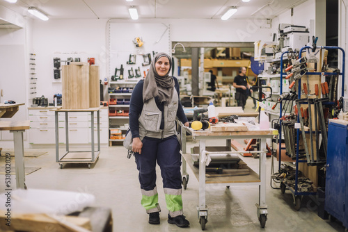 Full length portrait of smiling female carpenter standing by workbench in workshop