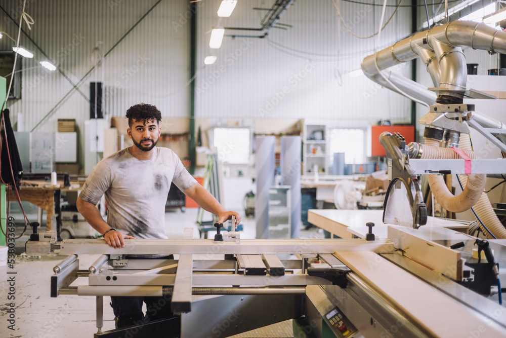Portrait of young carpenter standing by machinery in workshop Portrait of young carpenter standing by machinery in workshop