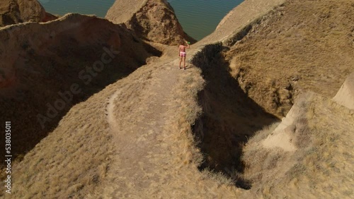 aerial footage of fitness woman jogger stands at early morning outdoor on endge of beautiful clay canyon on sea shore and drinks water. active leisure and exercising at summer sunrise. photo