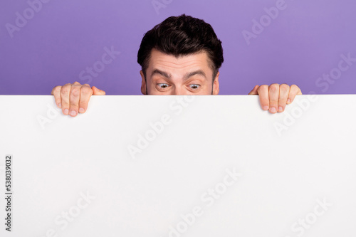 Photo portrait of nice young man playing hide and seek behind white poster look blank space isolated on purple color background