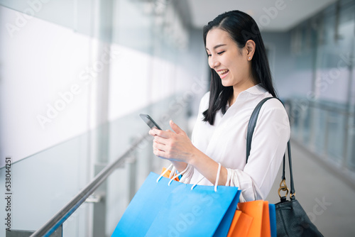 Happy Asian pretty girl holding shopping bags while using smartphone background shopping mall concept.