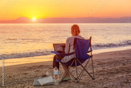 Girl with a laptop works online remotely on the coast of a sandy beach during a beautiful sunset.