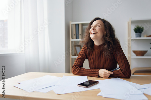 Enjoyed laughing happy cheerful curly cute lady sitting at table with papers documents work from home think about future holidays dream about weekend looks aside at window. Copy space