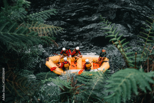Aerial view of five people wearing swimsuits and life jackets with oars in hand inside an inflatable boat ready to go rafting on the strong current of the river in Okere, new zealand