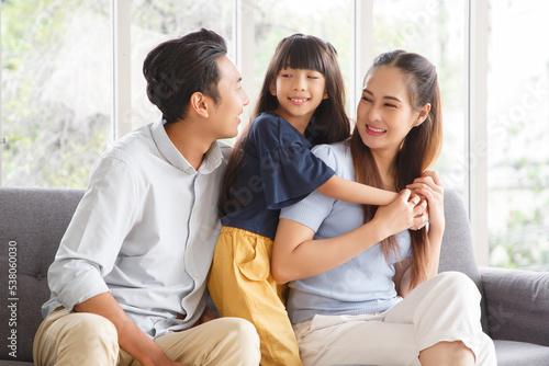 Portrait of a happy young family. Mom, dad and daughter look at the camera and smile. The faces of Asian parents and their child in living room.
