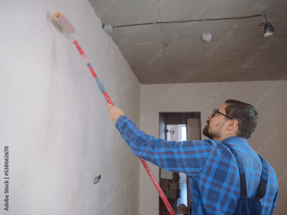 young man in blue work suit hold paint roller, over white wall room background. Instruments accessories for renovation apartment room. Repair home concept young man in blue work suit hold paint roller, over white wall room background. Instruments accessories for renovation apartment room. Repair home concept