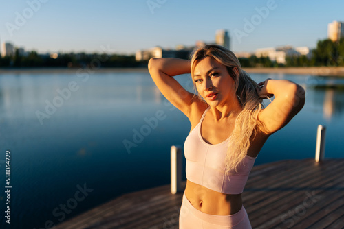 Portrait of beautiful sporty blonde woman in sportswear standing posing on wooden pier in cite park on sunny summer morning, looking at camera. Concept of outdoor sport and healthy lifestyle. © dikushin