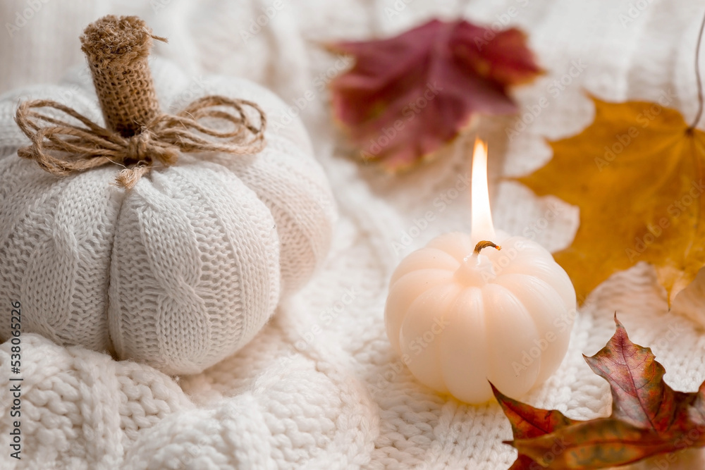 Still-life. Knitted pumpkin, autumn leaves, white knitted plaid and a burning candle in the shape of a pumpkin. Cozy autumn concept.