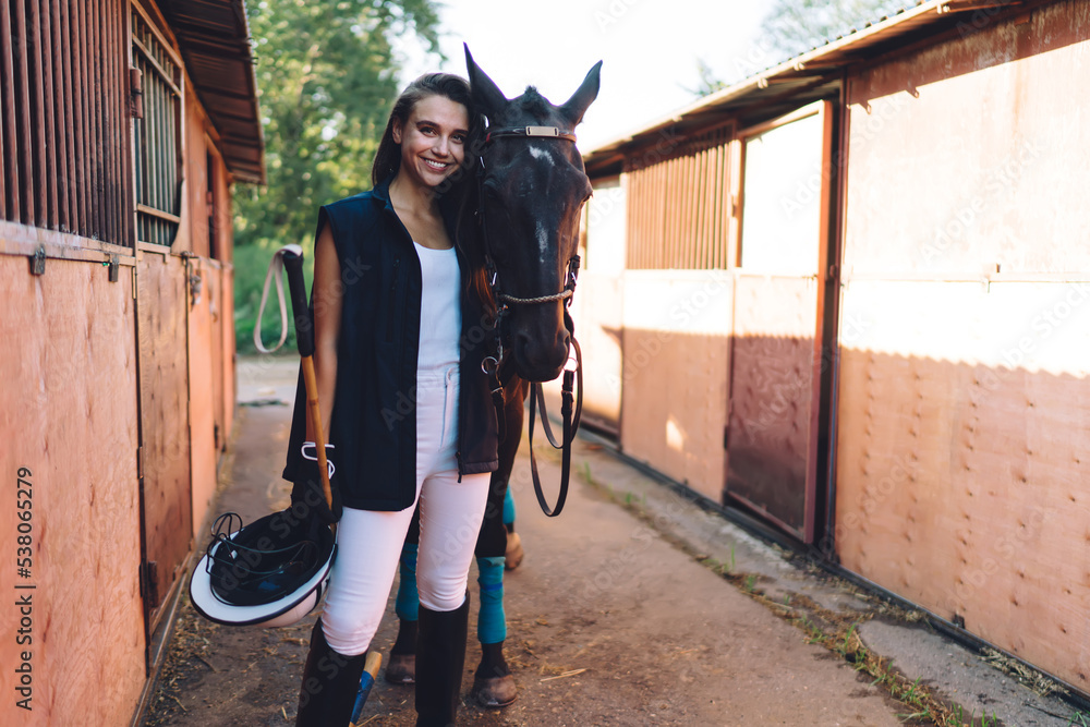 Naklejka premium Positive horsewoman embracing stallion near stables in countryside