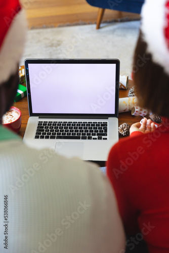 Happy diverse couple wearing santa claus hats, using laptop with copy space
