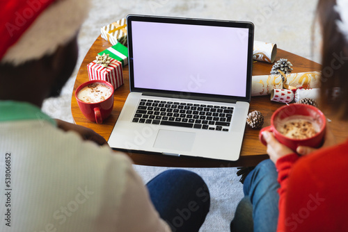 Happy diverse couple wearing santa claus hats, using laptop with copy space