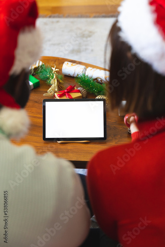 Happy diverse couple wearing santa claus hats, using tablet with copy space