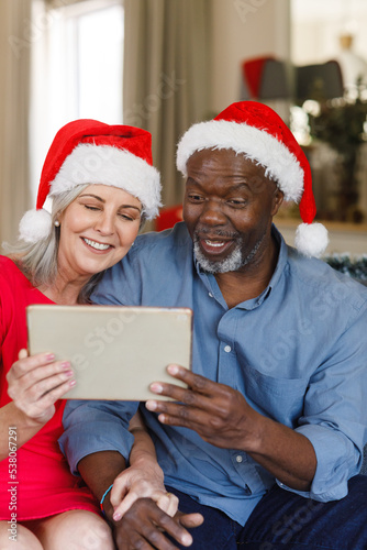 Happy senior diverse couple with santa hats using tablet