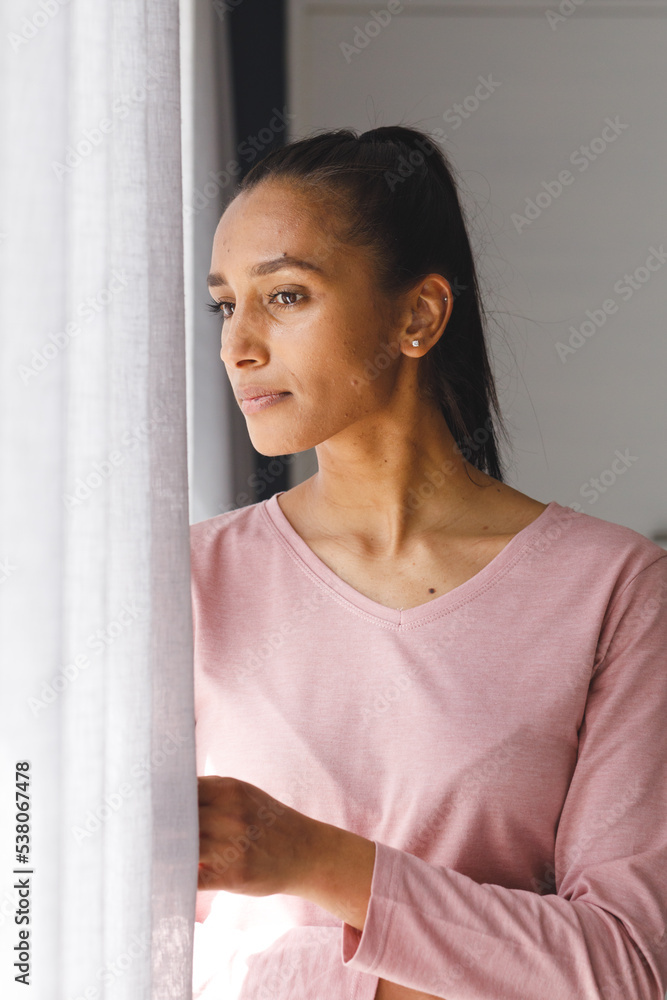 Vertical image of biracial woman looking through curtains out of window at home Vertical image of biracial woman looking through curtains out of window at home