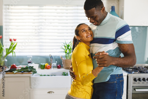 Happy diverse couple having fun slow dancing and smiling at each other in kitchen