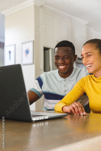 Vertical image of happy diverse couple making video call on laptop smiling at screen in kitchen