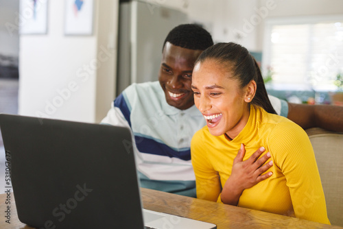 Happy diverse couple making video call on laptop smiling and laughing at screen in kitchen