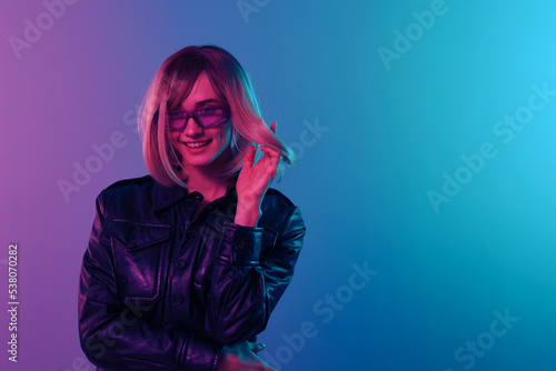 Flirting sexy smiling beautiful blonde woman in leather jacket sparkly dress trendy sunglasses posing isolated in blue pink color light studio background. Neon party Cyberpunk concept. Copy space