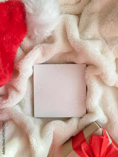 Next to a white empty square for text on a furry white background are a Santa Claus hat, a gift with a red bow.A Christmas card with a place for text. Copy space. Top view. Flatlay