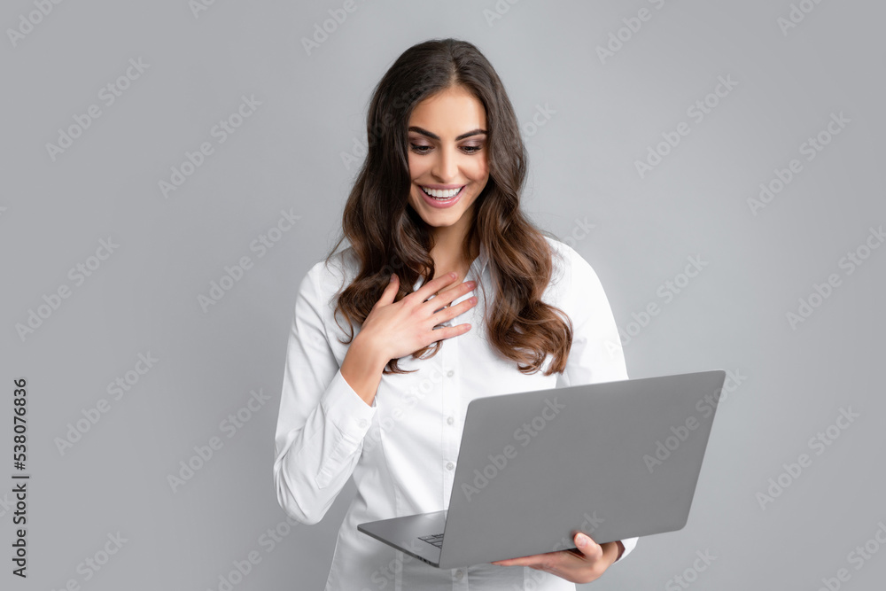 Cheerful business woman standing over grey wall with laptop computer. Portrait of pretty, charming, stylish, clever woman with open laptop chatting, using internet, isolated on grey background. Cheerful business woman standing over grey wall with laptop computer. Portrait of pretty, charming, stylish, clever woman with open laptop chatting, using internet, isolated on grey background.