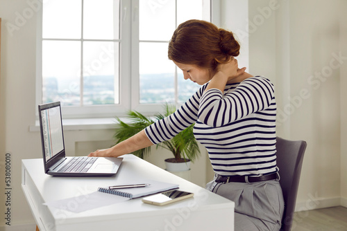 Very tired stressed young business woman sitting at office desk, holding hand on nape of her neck and feeling intense neck pain after working on laptop computer in uncomfortable position for long time