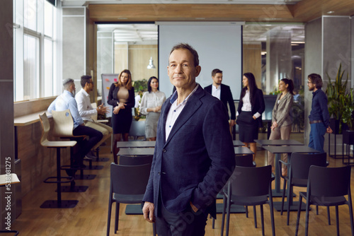 Successful adult Caucasian man manager posing in meeting room in front of business partners and employees company demonstrating confidence in future company, dressed in formal suit. Selective focus