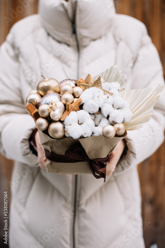 Very nice young woman holding humble and beautiful festive composition of cotton, ornaments, decorative apples, coconut stars in golden colors, cropped photo, bouquet close up