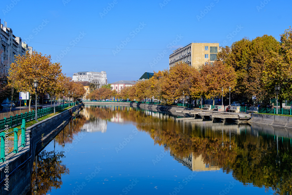 Fototapeta premium Landscape with Dambovita river, old buildings and yellow, orange and brown leaves in large trees in the center of Bucharest, Romania, in a sunny autumn day.