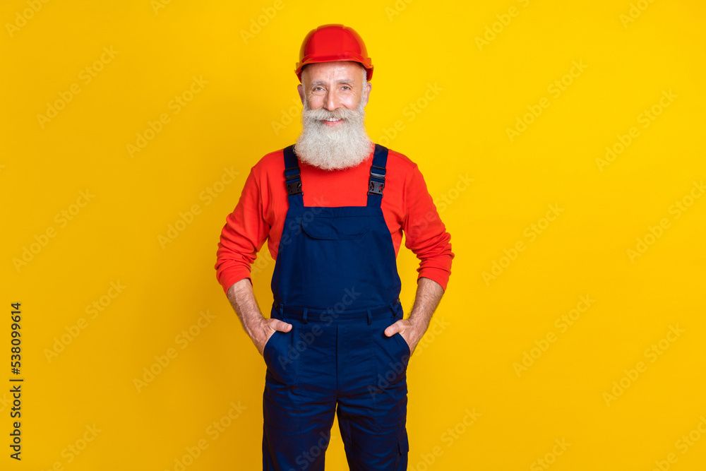 Photo of good mood cheerful senior guy dressed uniform overall red hardhat arms pockets isolated yellow color background