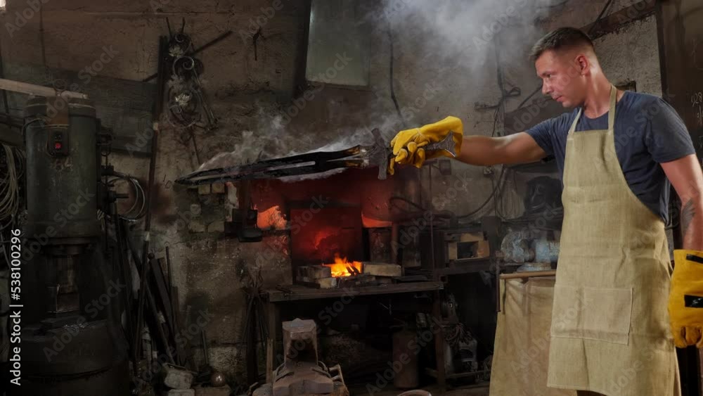 A male blacksmith holds a red-hot metal sword from which smoke is coming in an ancient forge. A male blacksmith holds a red-hot metal sword from which smoke is coming in an ancient forge.
